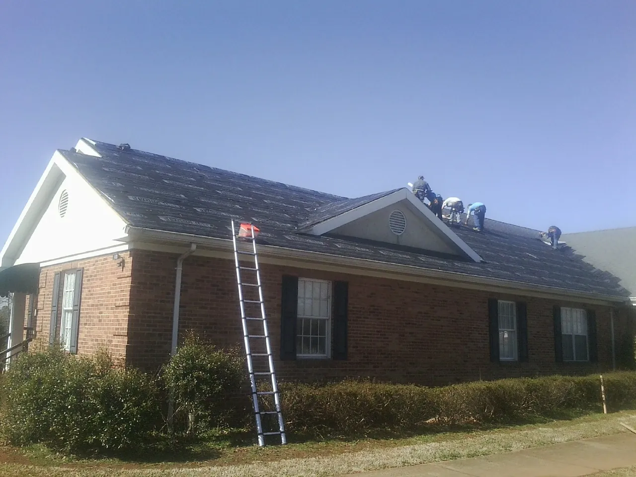 Crew tearing shingles off a brick ranch roof with a ladder set — Emergency Roof Repair work by our crew in Amery