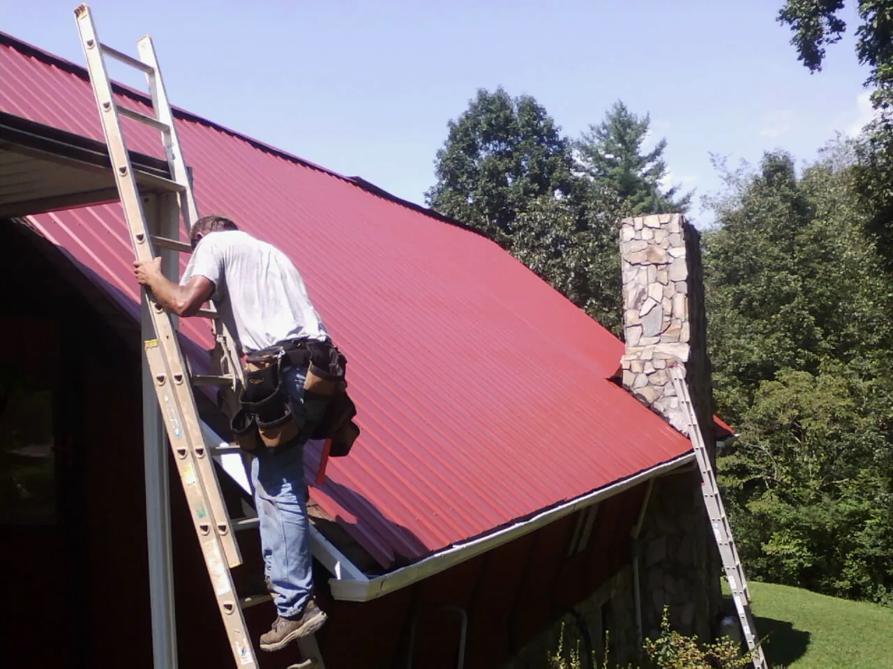 Roofer on a ladder beside a red metal roof and stone chimney — Soffit & Fascia Repair by Roofing Brothers in Amery